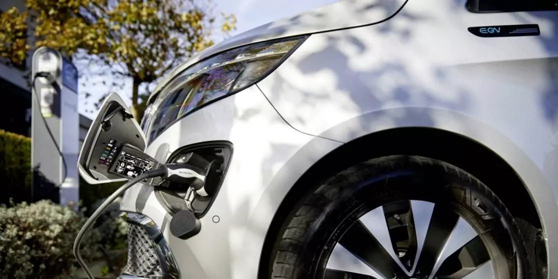 Wei&szlig;es Auto, an dessen vorderer Seitenbereich ein Ladekabel in die Ladebuchse gesteckt ist; vorderes Rad und Felge sichtbar; Lades&auml;ule und Baumschatten im Hintergrund