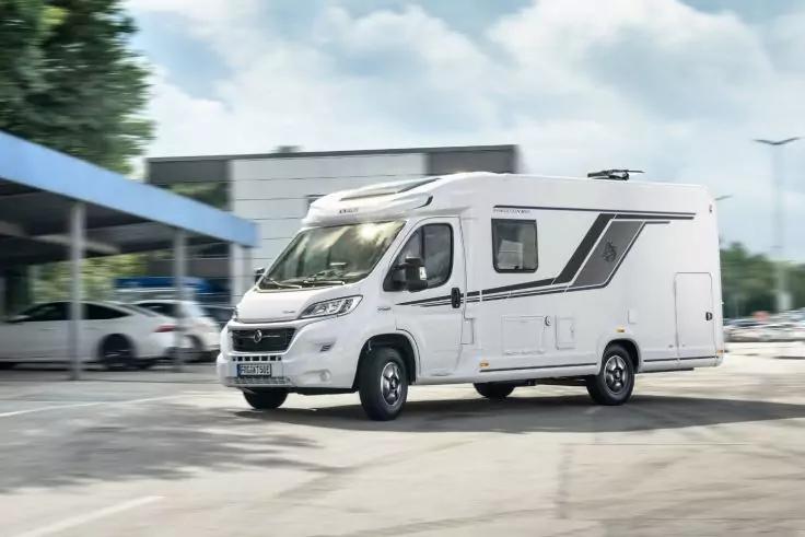 Wei&szlig;es Wohnmobil auf Asphalt vor einem Geb&auml;ude; links &uuml;berdachte Parkpl&auml;tze mit mehreren Autos; B&auml;ume und bew&ouml;lkter Himmel