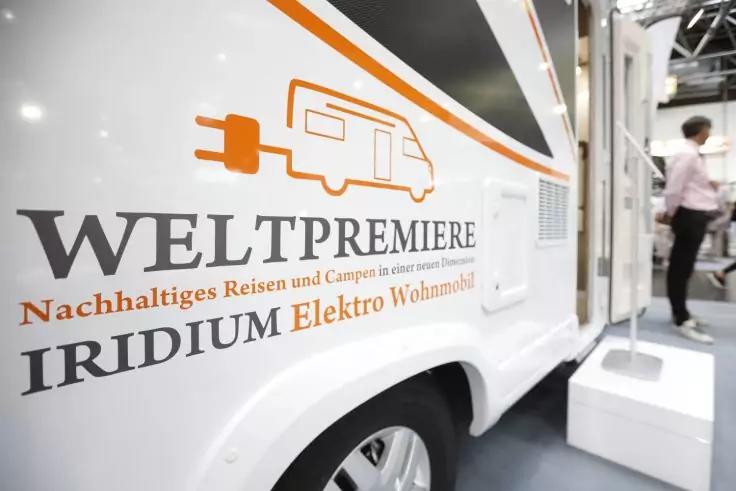 Wei&szlig;e Seitenansicht eines Wohnmobils mit orangefarbenem Steckersymbol und den Schriftz&uuml;gen 'WELTPREMIERE' und 'IRIDIUM Elektro Wohnmobil'; Person im Hintergrund
