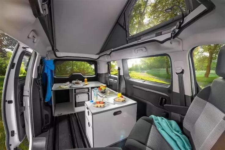 Innenraum eines Fahrzeugs mit offener Seitent&uuml;r, K&uuml;chenblock mit Sp&uuml;le und kleinem K&uuml;hlschrank, Arbeitsfl&auml;che mit Tellern, Gl&auml;sern und Obst, Sitze, ge&ouml;ffnete Dachluke und Fenster mit Blick auf B&auml;ume und Rasen