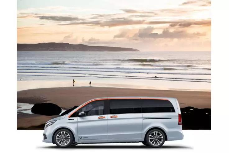 Silberner Kleinbus vor einem Sandstrand mit Wellen, drei Personen am Ufer, entfernte Klippen und wolkiger Himmel mit orange-rosa T&ouml;nen