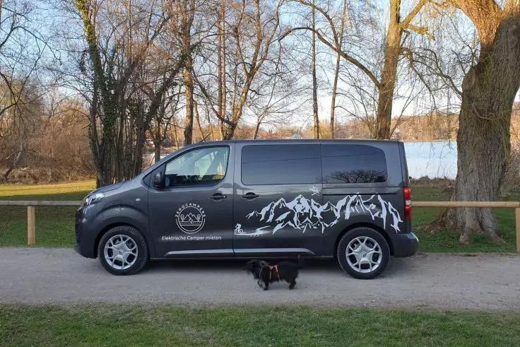 Seitliche Ansicht eines grauen Transporters mit wei&szlig;em Bergmotiv und Logo, geparkt auf einem Weg; davor liegt ein kleiner schwarzer Hund; im Hintergrund B&auml;ume, Rasen und ein See