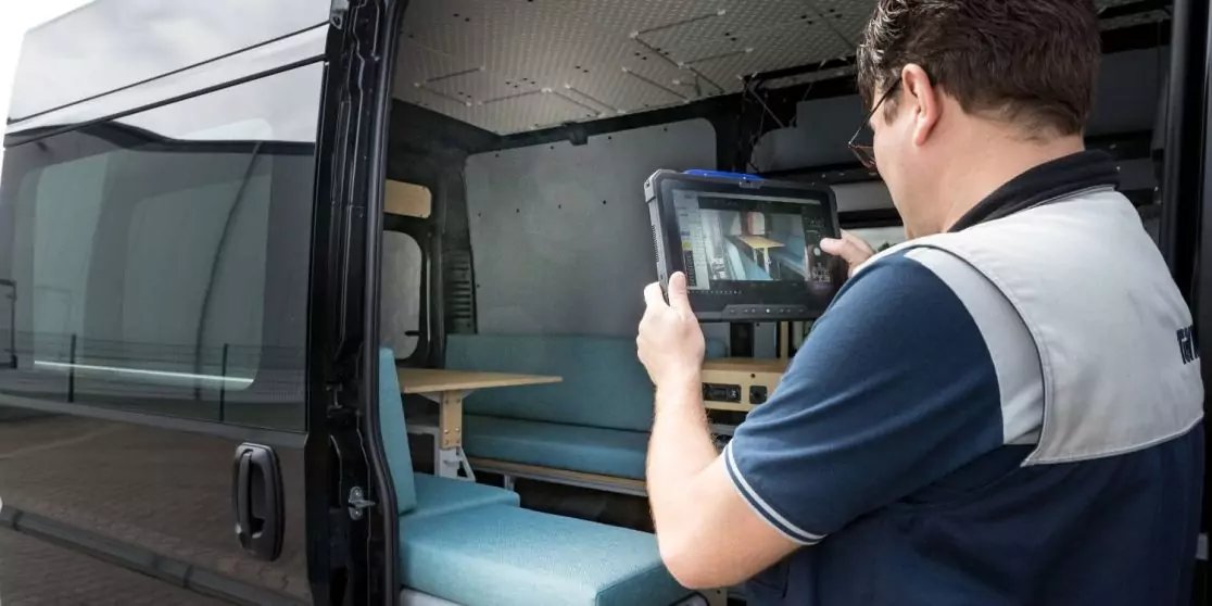 Ein Mann in Arbeitsweste steht in einem ge&ouml;ffneten Transporter und h&auml;lt ein Tablet; im Innenraum sind eine Sitzbank und Regale sichtbar