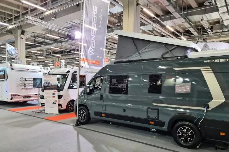 Dunkelgraues Wohnmobil mit aufgestelltem Hubdach in einer Messehalle, daneben wei&szlig;e Wohnmobile, Deckenbeleuchtung und h&auml;ngende Messebanner