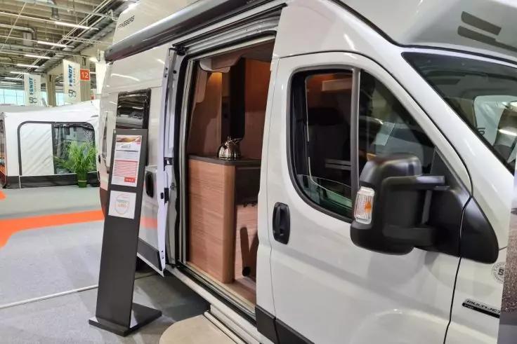 Wei&szlig;es Wohnmobil mit ge&ouml;ffneter Seitent&uuml;r, sichtbare K&uuml;chenzeile und Sitzbank im Innenraum, schwarzer Displayst&auml;nder neben der T&uuml;r, weiterer Wohnwagen und Halleninnenraum im Hintergrund