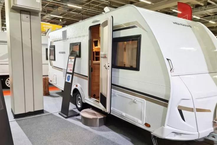 Wei&szlig;er Wohnwagen in einer Halle, ge&ouml;ffnete T&uuml;r mit kleinem Tritt, Informationsaufsteller neben der T&uuml;r