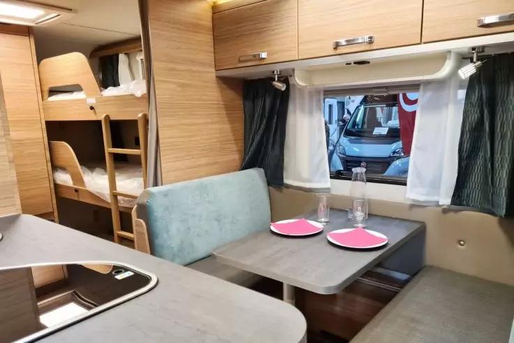 Innenraum eines Wohnmobils: Tisch mit zwei roten Tellern und zwei Gl&auml;sern, Sitzb&auml;nke, Etagenbetten mit Leiter, Holzschr&auml;nke, K&uuml;chenarbeitsplatte und Fenster mit Vorh&auml;ngen