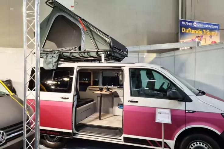 Wei&szlig;-rosa Transporter mit aufgestelltem Dachkoffer und offener Schiebet&uuml;r, Innenraum mit Tisch sichtbar, Messestand im Hintergrund