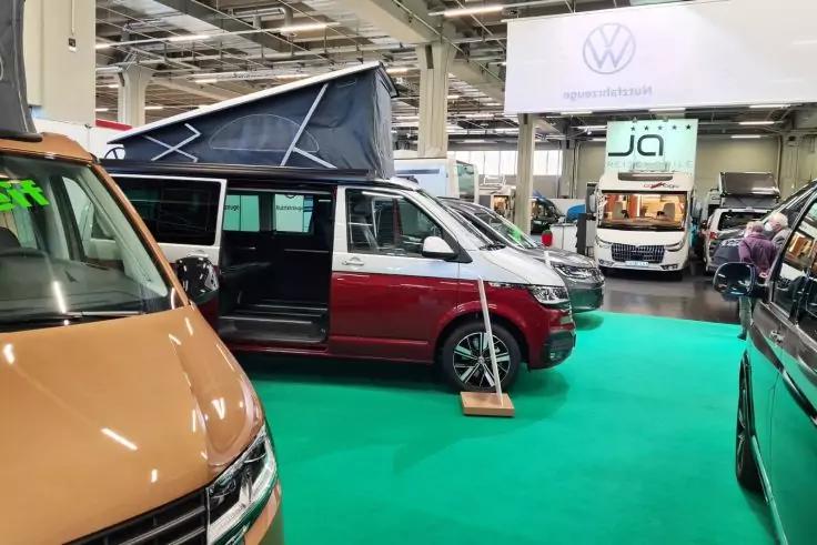 Innenaufnahme mehrerer Campingvans in einer Halle, gr&uuml;ner Bodenbelag, wei&szlig;es Schild mit VW-Logo, rot-wei&szlig;es Fahrzeug mit ge&ouml;ffneter Schiebet&uuml;r und aufgestelltem Hochdach, weitere Vans im Hintergrund