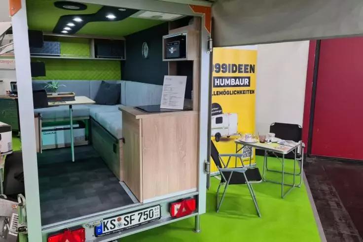 Offener Anh&auml;nger mit eingebauter Sitzecke, Tisch und Schr&auml;nken; rechts gelbes Banner und Klappst&uuml;hle auf gr&uuml;nem Boden