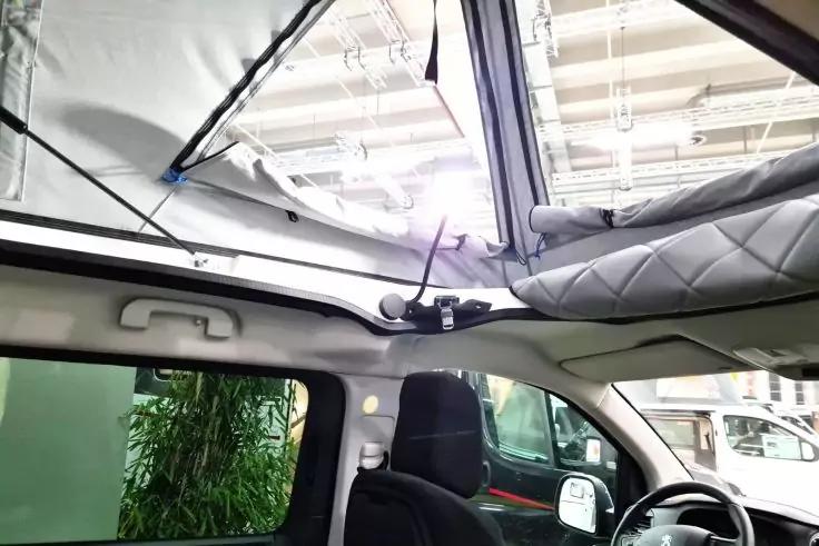 Innenraum eines Fahrzeugs mit ge&ouml;ffnetem Aufstelldach, vorderen Sitzen, Lenkrad, Armaturenbrett und ge&ouml;ffneter Seitenklappe; im Hintergrund weiteres Fahrzeug und Pflanzen