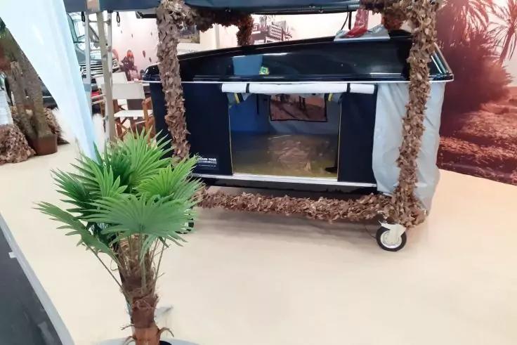 Rollwagen mit Rahmen, der mit braunen Fasern umwickelt ist; auf dem Wagen eine schwarze Box mit offenem Fach; vorne links eine kleine k&uuml;nstliche Palmenpflanze auf hellem Boden