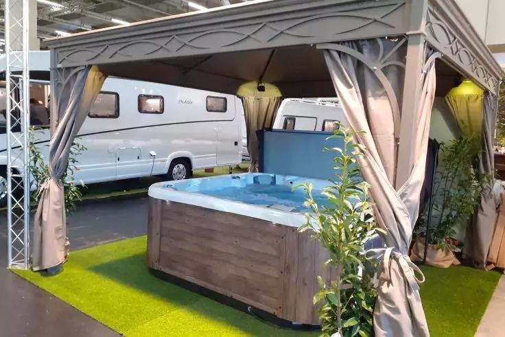 Whirlpool unter einem Pavillon mit ge&ouml;ffneten Vorh&auml;ngen auf Kunstgras, umgeben von Pflanzen; Wohnwagen im Hintergrund