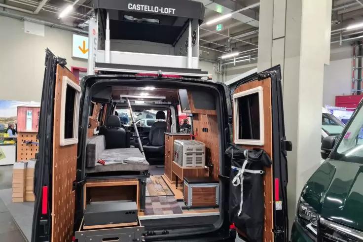 Offene Heckt&uuml;ren eines schwarzen Vans in einer Messehalle; sichtbarer Innenraum mit Holzverkleidung, Schubladen, Arbeitsfl&auml;che mit Sp&uuml;le und Kochfeld, Liegefl&auml;che, Regale und Deckenspots; Schild "CASTELLO-LOFT" &uuml;ber dem Fahrzeug
