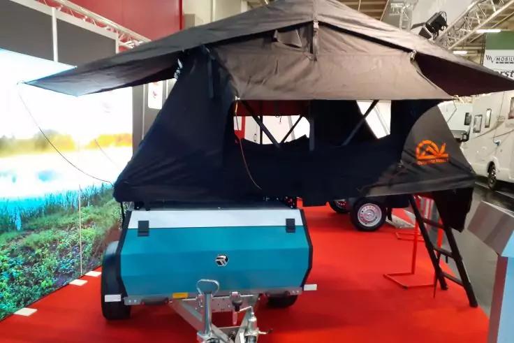 T&uuml;rkisfarbener Anh&auml;nger mit aufgeklapptem schwarzem Dachzelt und Leiter auf rotem Teppich in Halle