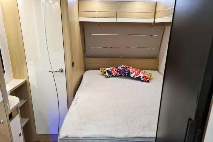 Bett mit wei&szlig;er Decke und buntem Blumen-Kissen; Holzschr&auml;nke &uuml;ber dem Kopfende; geschlossene T&uuml;r links; kleines Waschbecken und Regalf&auml;cher sichtbar