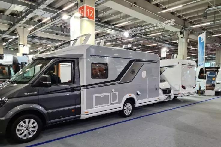 Graues Wohnmobil vorne links, wei&szlig;e Wohnmobile dahinter in gro&szlig;er Halle; S&auml;ule mit orangefarbigem Schild "12", Deckenlampen und blaue Bodenmarkierung