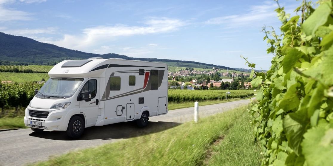 Wei&szlig;es Wohnmobil auf einer Landstra&szlig;e neben Weinreben, Dorf und H&uuml;gel im Hintergrund, blauer Himmel