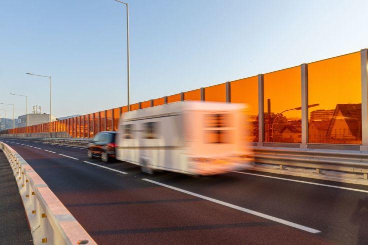 Autobahn mit Leitplanke links, orangefarbene Schallschutzwand rechts, wei&szlig;er Wohnwagen und dunkles Auto mit Bewegungsunsch&auml;rfe, Stra&szlig;enlaternen und Himmel