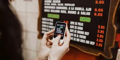 Zwei H&auml;nde halten ein Smartphone mit sichtbarem Bildschirm; im Hintergrund eine Men&uuml;tafel mit Speisen und Preisen