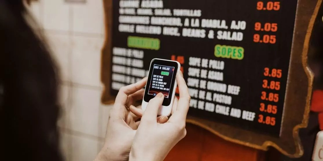 Zwei H&auml;nde halten ein Smartphone mit sichtbarem Bildschirm; im Hintergrund eine Men&uuml;tafel mit Speisen und Preisen