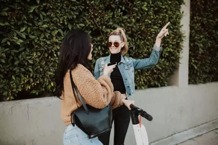 Zwei Frauen auf einem B&uuml;rgersteig vor einer Hecke. Links: hellblaue Jeans, beiger Strickpullover, schwarze Umh&auml;ngetasche, h&auml;lt ein Smartphone. Rechts: Jeansjacke, schwarze Kleidung, Sonnenbrille, steht neben einem E‑Scooter, hebt den rechten Arm und