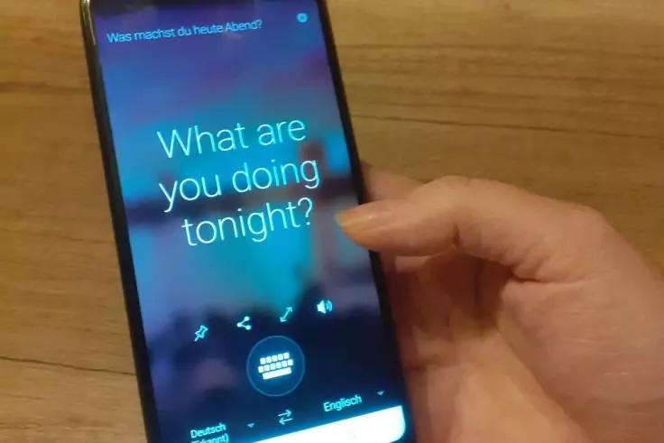Hand h&auml;lt ein Smartphone &uuml;ber einer Holzoberfl&auml;che, ein Finger ber&uuml;hrt den Bildschirm; auf dem Display steht 'What are you doing tonight?' und oben 'Was machst du heute Abend?'