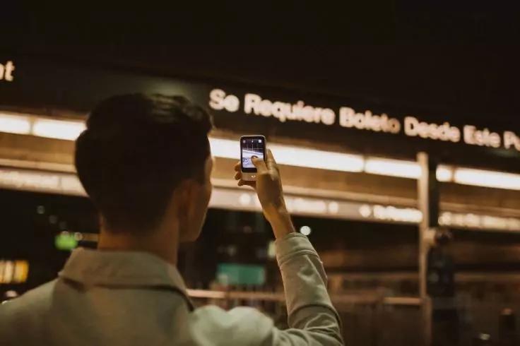 Person von hinten h&auml;lt ein Smartphone hoch; im Hintergrund leuchtende Anzeigetafel mit Text