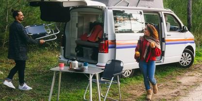 Wei&szlig;er Campingbus mit offener Seitent&uuml;r auf einem Waldweg; davor zwei Personen, links h&auml;lt eine zusammengeklappte Sitzgelegenheit, rechts steht eine Person mit einer Tasse; vor dem Bus ein Campingtisch mit Wasserkocher, Tasse und Campingstuhl