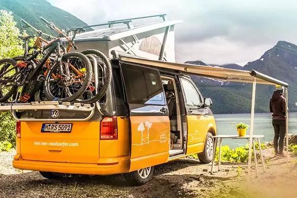 Gelb-orangener Campingbus mit aufgeklapptem Dachzelt, Fahrradträger mit mehreren Fahrrädern am Heck, ausgefahrene Markise, kleiner Tisch und Stuhl, stehende Person mit dem Rücken zur Kamera Richtung See, Berge im Hintergrund