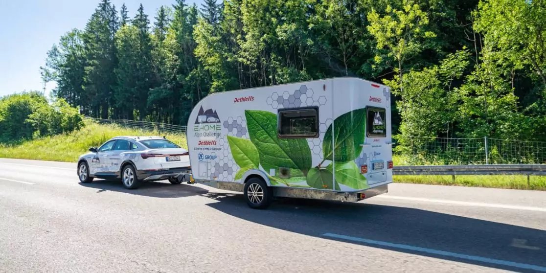Silbernes Auto mit wei&szlig;em Wohnwagen mit gr&uuml;nen Blattmotiven auf einer Stra&szlig;e, B&auml;ume und Gras im Hintergrund, sonniger Himmel