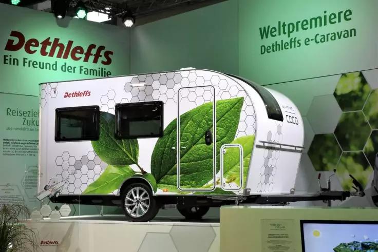 Wei&szlig;er Caravan mit gro&szlig;em gr&uuml;nen Blatt-Design und Dethleffs-Logo auf einer Ausstellungsplattform, im Hintergrund die Schriftz&uuml;ge 'Weltpremiere' und 'Dethleffs e-Caravan'