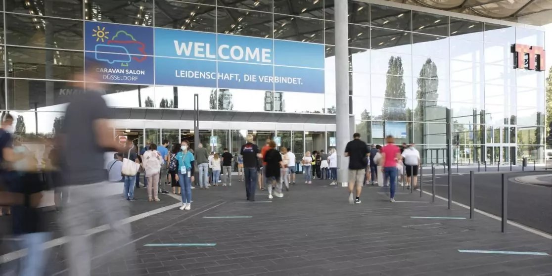 Eingangsbereich eines Geb&auml;udes mit Glasfassade und gro&szlig;em Banner 'WELCOME' &uuml;ber den T&uuml;ren. Mehrere Personen gehen zum Eingang und stehen an Absperrungen