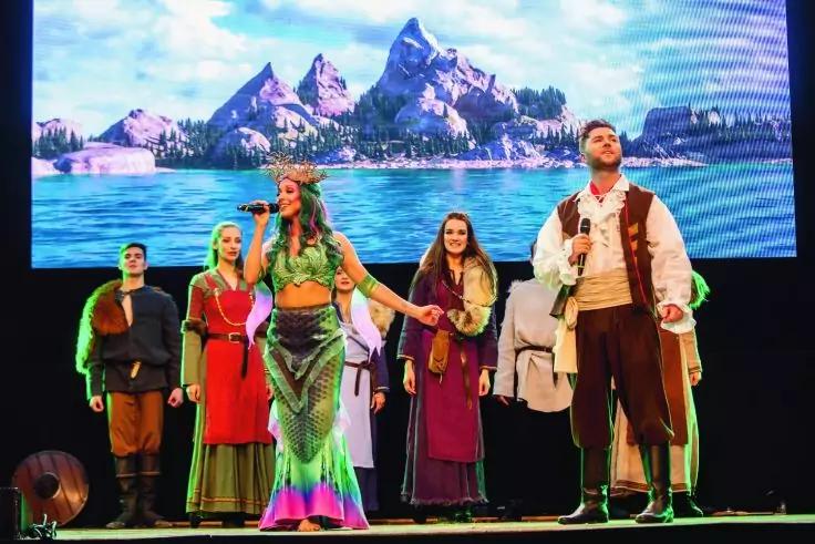 Mehrere kost&uuml;mierte Darsteller auf einer B&uuml;hne; vorn eine Frau in gr&uuml;nem, schuppenartigem Kost&uuml;m mit flossen&auml;hnlichem Saum, daneben ein Mann mit Mikrofon; im Hintergrund ein gro&szlig;es Bildschirmbild von Meer und felsigen Inseln