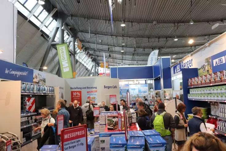 Innenaufnahme einer Messehalle mit mehreren St&auml;nden, Regalen und Personen, die sich zwischen den G&auml;ngen bewegen