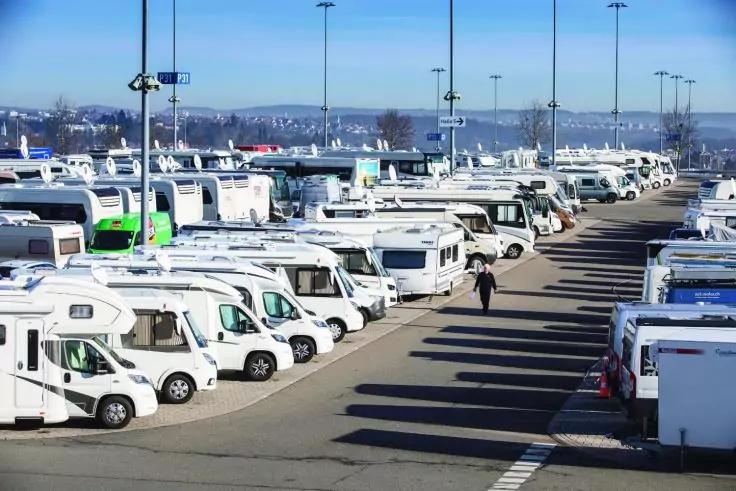 Viele wei&szlig;e Wohnmobile in Reihen auf einem gepflasterten Parkplatz, eine Person geht dazwischen, hohe Laternen, Geb&auml;ude und H&uuml;gel im Hintergrund, blauer Himmel
