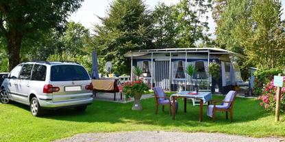 Wohnwagen mit angebautem Vorzelt, davor ein Tisch mit zwei St&uuml;hlen und Blument&ouml;pfen auf Rasen, links ein silberner Van, B&auml;ume im Hintergrund