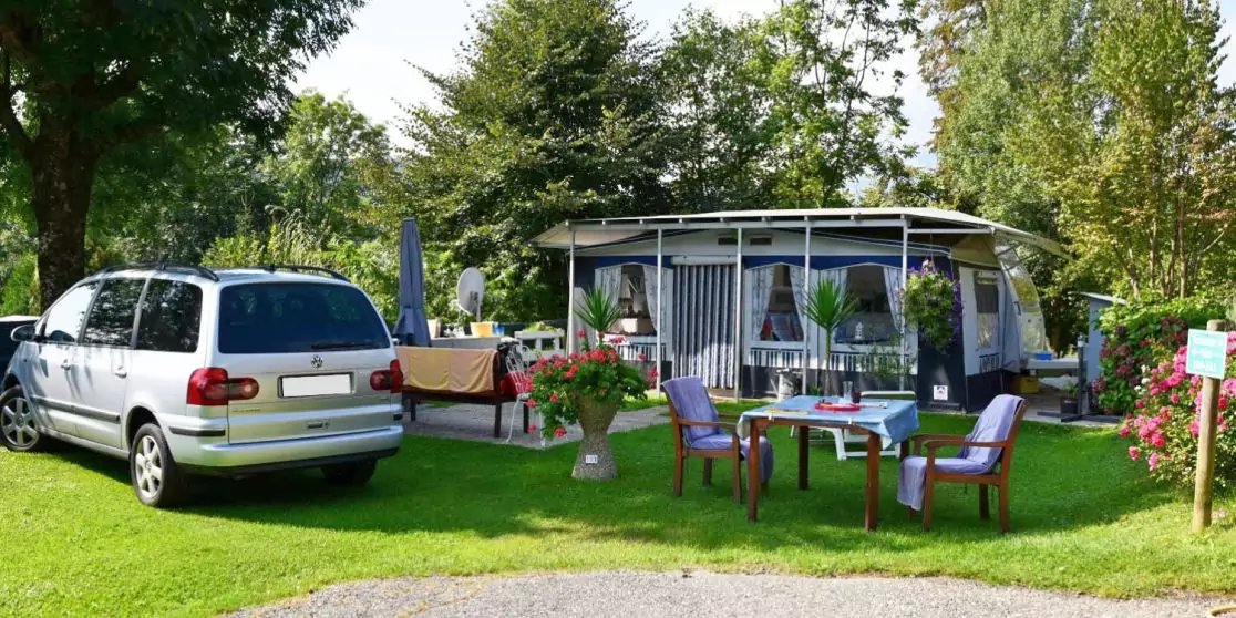 Wohnwagen mit angebautem Vorzelt, davor ein Tisch mit zwei St&uuml;hlen und Blument&ouml;pfen auf Rasen, links ein silberner Van, B&auml;ume im Hintergrund