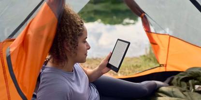 Person liegt in der &Ouml;ffnung eines orangefarbenen Zelts und h&auml;lt einen E-Reader; See und Gras im Hintergrund