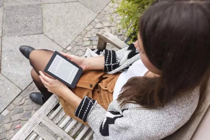 Person sitzt auf einer Holzbank und h&auml;lt einen E-Reader mit sichtbarem Text, tr&auml;gt grauen Cardigan und braunen Rock