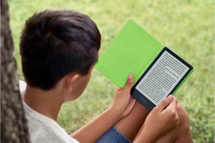 Person von hinten sitzt auf einer Wiese und h&auml;lt einen E-Reader mit gr&uuml;nem Cover; auf dem Display ist Text sichtbar