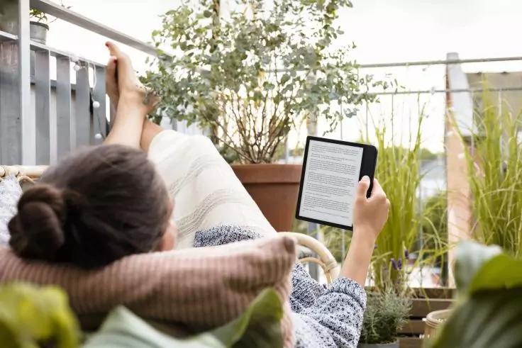 Person von hinten auf einer gepolsterten Liege auf einem Balkon liegend, h&auml;lt ein E-Reader mit sichtbarem Text; Beine auf dem Gel&auml;nder; Pflanzen in T&ouml;pfen umgeben sie