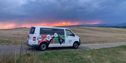 Wei&szlig;er Lieferwagen mit buntem Werbeaufdruck auf einer schmalen Landstra&szlig;e, abgeerntete Felder daneben, H&uuml;gel im Hintergrund, bew&ouml;lkter Himmel und orangefarbener Sonnenuntergang