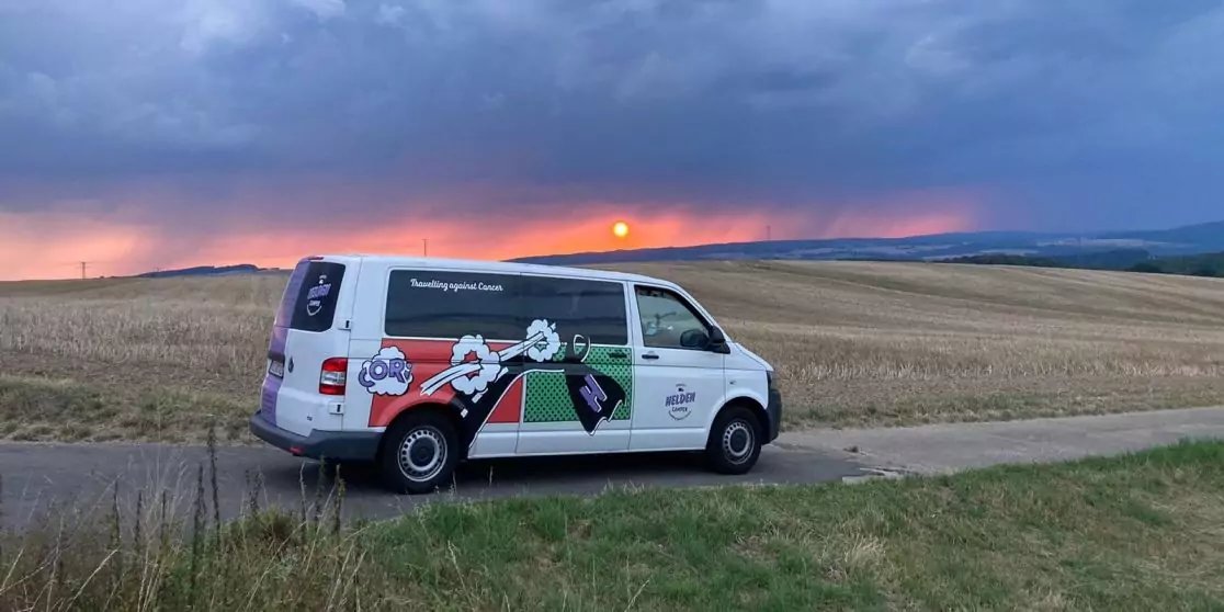 Wei&szlig;er Lieferwagen mit buntem Werbeaufdruck auf einer schmalen Landstra&szlig;e, abgeerntete Felder daneben, H&uuml;gel im Hintergrund, bew&ouml;lkter Himmel und orangefarbener Sonnenuntergang