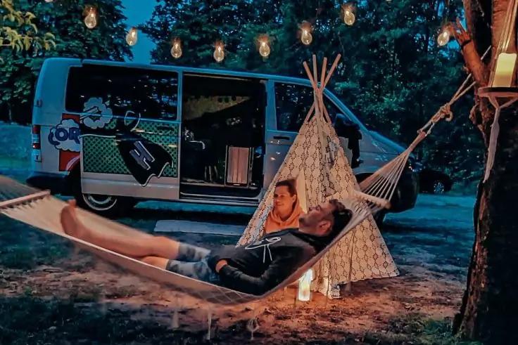 Zwei Personen liegen in einer H&auml;ngematte unter aufgeh&auml;ngten Lichterketten neben einem offenen Wohnmobil und einem kleinen Tipi, eine Laterne steht im Vordergrund, dunkle Au&szlig;enaufnahme mit B&auml;umen