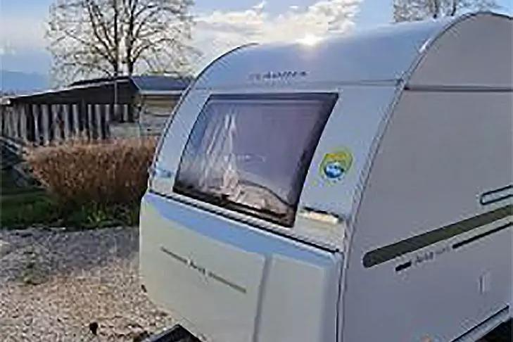 Wei&szlig;er Caravan mit Fenster und Aufkleber, geparkt auf Schotter vor einem kleinen Geb&auml;ude, Baum und Berge im Hintergrund