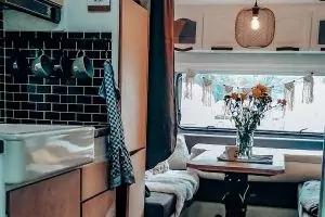 Innenraum mit K&uuml;chenzeile (Sp&uuml;le, schwarze Fliesenr&uuml;ckwand, h&auml;ngende Tassen), Tisch mit Sitzbank, Glasvase mit Blumen, Fenster mit Vorhang, h&auml;ngende Lampe