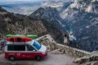 Roter Van mit zwei Kajaks auf dem Dach, zwei Personen auf einer steinernen Mauer, Gebirgsschlucht und Wasserfl&auml;che im Hintergrund