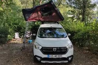 Wei&szlig;es Auto mit ge&ouml;ffnetem rotem Dachzelt und Leiter, B&auml;ume im Hintergrund, Laub auf dem Boden, klappbarer Stuhl neben dem Auto