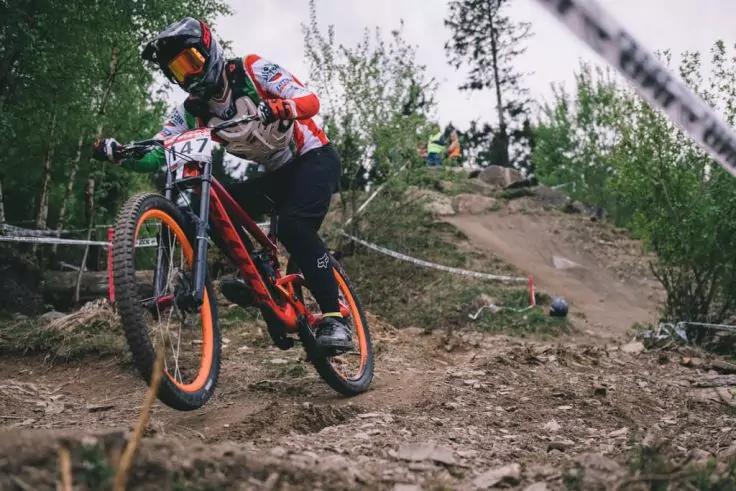 Person auf orangefarbenem Mountainbike mit Startnummer, tr&auml;gt Fullface-Helm und Schutzbrille, f&auml;hrt einen steinigen, mit Absperrband markierten Waldweg hinab